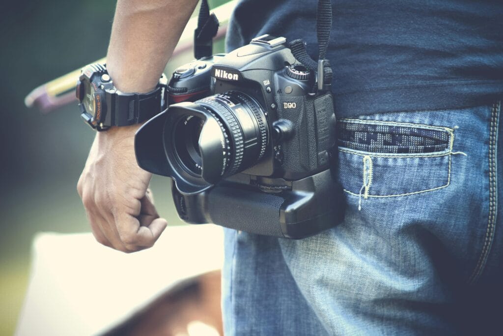 Gdzie można nas spotkać? A photographer holds a DSLR camera, wearing casual denim jeans and wristwatch outdoors.